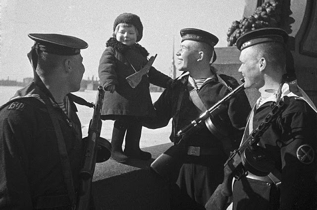 Leningrad—Despite the siege there must be some light hearted moments.  Here, sailors of the Baltic Fleet play with two year old Lucy, who had recently lost her parents in a bombardment. If Lucy is still alive she will be seventy years old. 